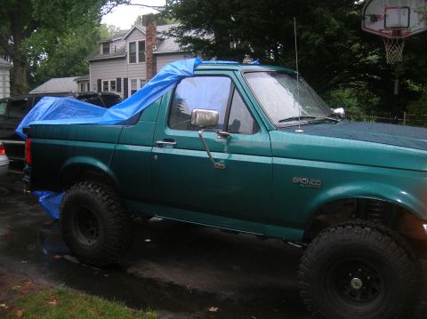 1996 Ford Bronco - Side view of a lifted teal-green Ford Bronco XLT sitting in a wet driveway with a blue tarp draped over the open rear cargo area in place o…