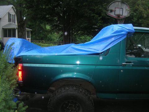 1996 Ford Bronco - A green SUV is partially covered by a blue tarp, parked outdoors near a house.