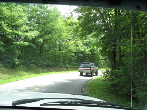 1995 Ford C-350 (Centurion Conversions) - Driving behind a Ford Bronco on a winding road surrounded by dense green trees.
