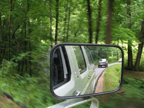 1995 Ford C-350 (Centurion Conversions) - A white pickup truck is seen in the side mirror of another vehicle as they drive down a tree-lined road.