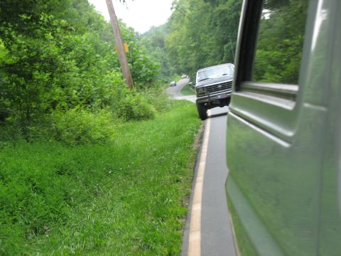 1995 Ford C-350 (Centurion Conversions) - A dark 1980s Ford F-Series truck navigates a curved road surrounded by lush green trees and foliage.