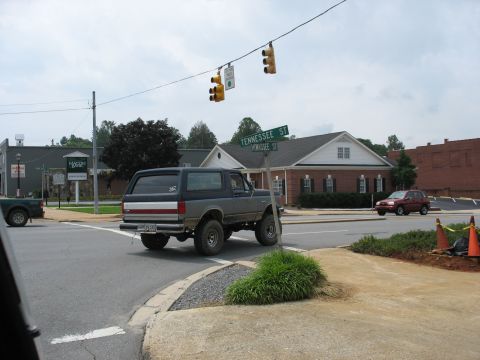 1995 Ford C-350 (Centurion Conversions) - A lifted gray Ford Bronco turns at an intersection, with a red SUV visible in the background.