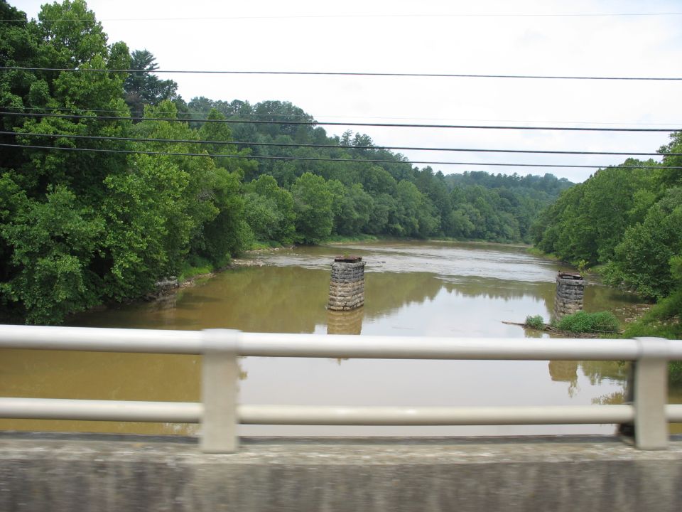 River flowing between tree-lined banks, with two stone bridge supports visible in the water and a white railing in the foreground