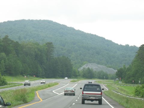 1995 Ford C-350 (Centurion Conversions) - A pickup truck travels down a highway, with a large pile of gravel visible near a forested mountain.