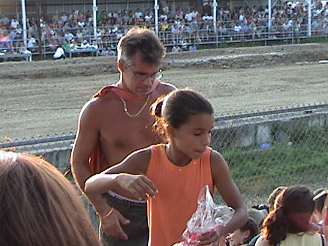 Man with a cape and a girl in an orange shirt at an outdoor event with spectators in the background