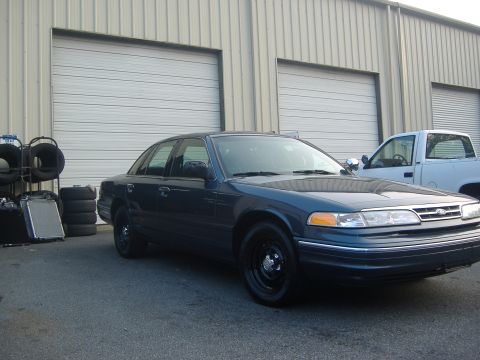 2001 Ford Crown Victoria - A dark blue sedan is parked outside a commercial building next to a white pickup truck, with spare tires and a radiator nearby.
