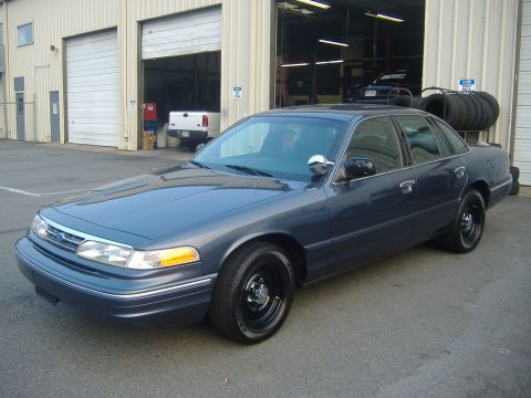 2001 Ford Crown Victoria - A blue 2001 Ford Crown Victoria sedan is parked in front of a garage with tires stacked on a rack behind it.