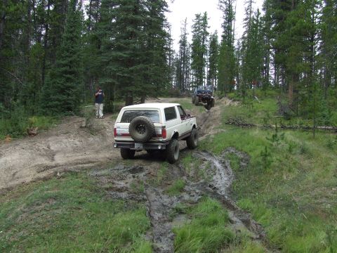1990 Ford Bronco - Two SUVs navigate a challenging, muddy off-road trail through a dense forest.