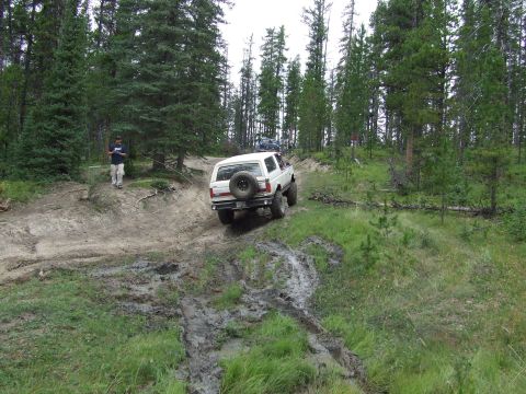 1990 Ford Bronco - A white Ford Bronco navigates a muddy track through a dense forest.
