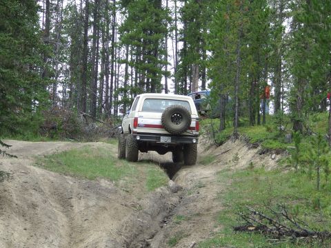 1990 Ford Bronco - A 1990 Ford Bronco navigates a challenging dirt path in a wooded area, with a second vehicle following.