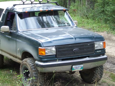 1990 Ford Bronco - A green Ford Bronco with off-road tires is parked on a dirt road surrounded by trees.