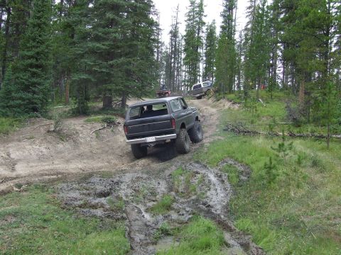 1990 Ford Bronco - A dark 1978 Ford Bronco navigates a muddy, wooded trail, with another vehicle following behind.