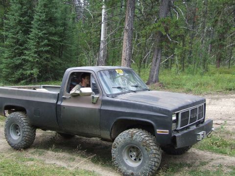 1990 Ford Bronco - A lifted pickup truck with aggressive off-road tires sits on a dirt path surrounded by trees.