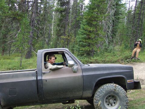 1990 Ford Bronco - A man drives a pickup truck through a wooded area while another person sits on a stump in the background.