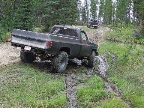 1990 Ford Bronco - Two lifted pickup trucks tackle a challenging muddy off-road course in a wooded area.