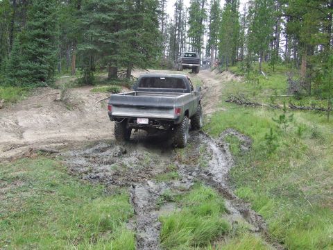 1990 Ford Bronco - Two pickup trucks navigate a muddy off-road trail surrounded by trees.