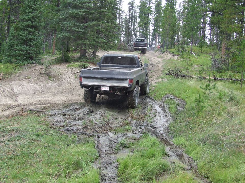 1973 Chevrolet C/K pickup truck driving through mud, another truck behind it on a dirt trail
