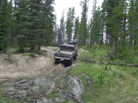 1990 Ford Bronco - Two trucks navigate a challenging, muddy off-road trail through a dense forest.