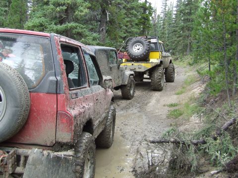 1990 Ford Bronco - Multiple off-road vehicles navigate a muddy path through a dense forest.