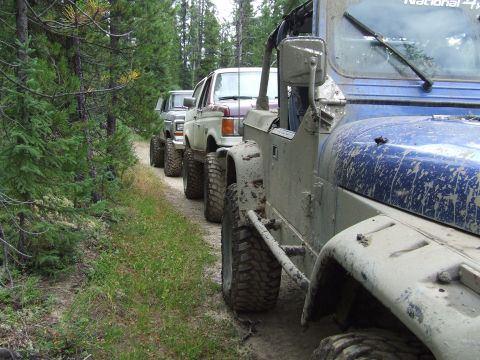 1990 Ford Bronco - A convoy of off-road vehicles navigates a dirt trail through a dense forest, kicking up dust and mud.