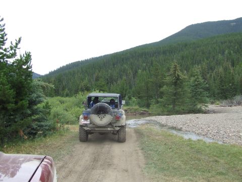 1990 Ford Bronco - A Jeep Wrangler navigates a dirt road through a forested mountain landscape.