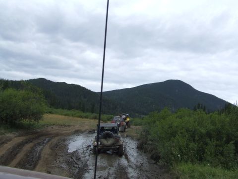 1990 Ford Bronco - Several off-road vehicles navigate a challenging muddy track through a wooded, hilly landscape.