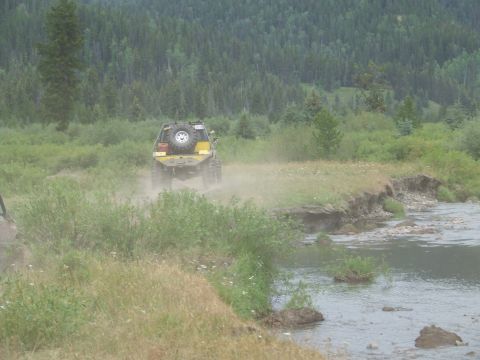 1990 Ford Bronco - A yellow 1990 Ford Bronco kicks up dust as it drives along a dirt path next to a shallow river.