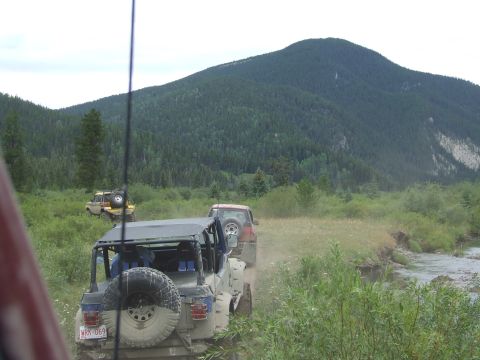 1990 Ford Bronco - Several off-road vehicles navigate a dusty trail alongside a flowing stream with a large, tree-covered mountain in the distance.