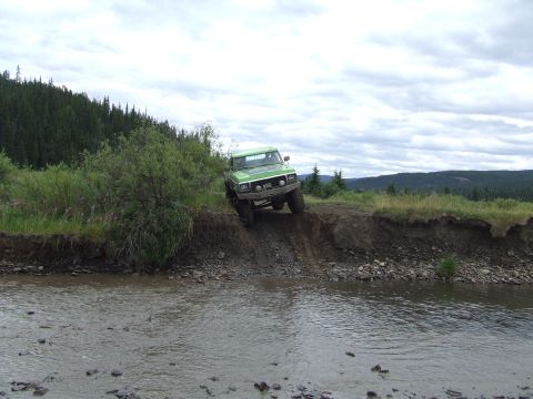 1990 Ford Bronco - A green off-road truck navigates a steep dirt bank next to a rocky stream under a cloudy sky.