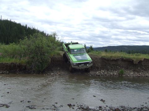 1990 Ford Bronco - A modified green pickup truck navigates a rocky stream bed with a forested hillside in the background.