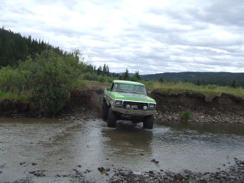 1990 Ford Bronco - A lifted green Ford F-Series pickup truck navigates a rocky stream bed under a cloudy sky.