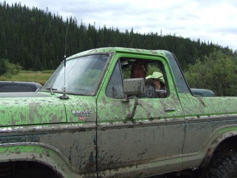 1990 Ford Bronco - A mud-covered Ford Bronco drives through a wooded area with two people inside.