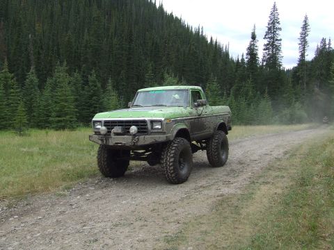 1990 Ford Bronco - A heavily modified green pickup truck drives on a dusty mountain road, with another vehicle in the distance.