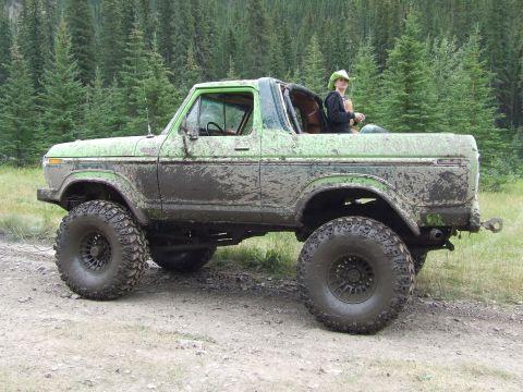 1990 Ford Bronco - A heavily muddied green Ford Bronco sits off-road with a person in a cowboy hat visible in the passenger seat.