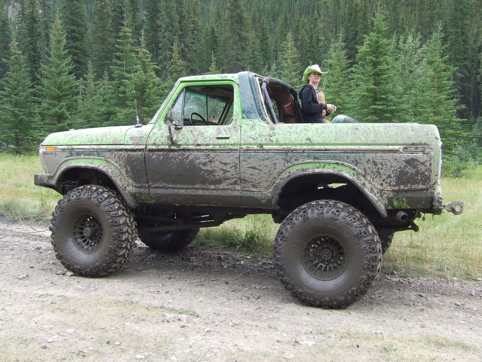 Mud-covered green Ford Bronco with large tires, person in cowboy hat in passenger seat