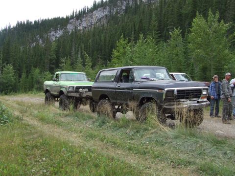1990 Ford Bronco - Two lifted 4x4 trucks, a black Bronco and a green pickup, are parked on a dirt road in a wooded mountain area.