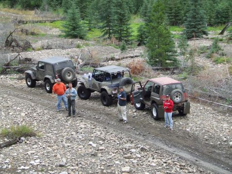 1990 Ford Bronco - Several off-road vehicles and people are gathered on a gravel path in a wooded area.