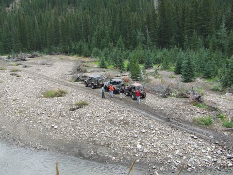 1990 Ford Bronco - A group of off-road vehicles are stopped on a gravel road near a river, surrounded by dense pine forest.