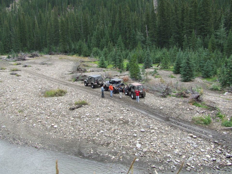 Several Jeeps parked on a rocky dirt road with people standing around them. Forested mountains are in the background