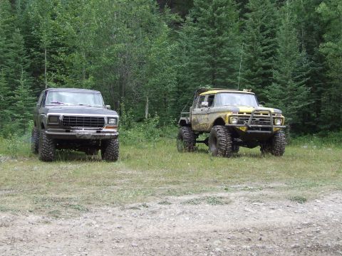 1990 Ford Bronco - Two modified Ford trucks, a Bronco and an F-series, are parked off-road in a wooded area.