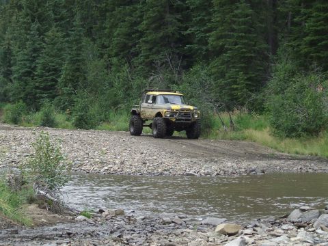1990 Ford Bronco - Modified yellow pickup truck navigates a rocky stream crossing on a forest road.