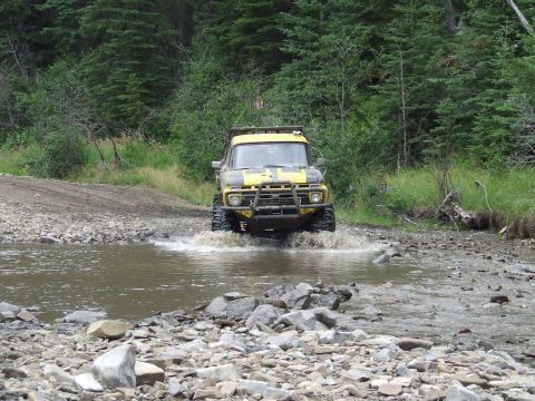 1990 Ford Bronco - An off-road truck navigates a rocky stream bed surrounded by trees.