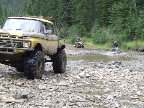 1990 Ford Bronco - A yellow off-road truck navigates a rocky stream with ATVs and motorcycles in the background.