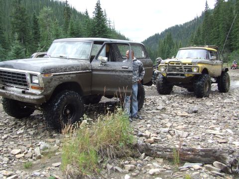 1990 Ford Bronco - Two off-road vehicles are parked on a rocky riverbed surrounded by a forest.