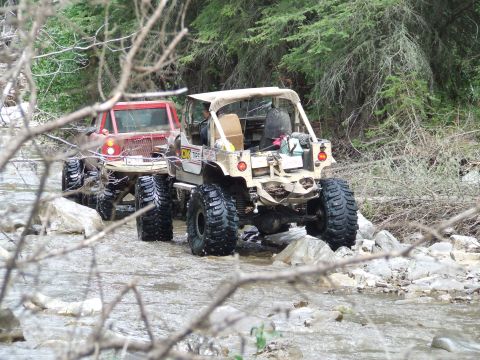 1990 Ford Bronco - Off-road vehicles traverse a shallow, rocky stream surrounded by trees.