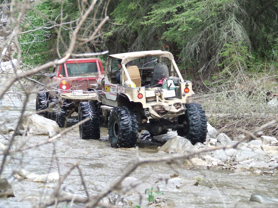 Two off-road vehicles with large tires navigate a rocky stream, one tan and one red