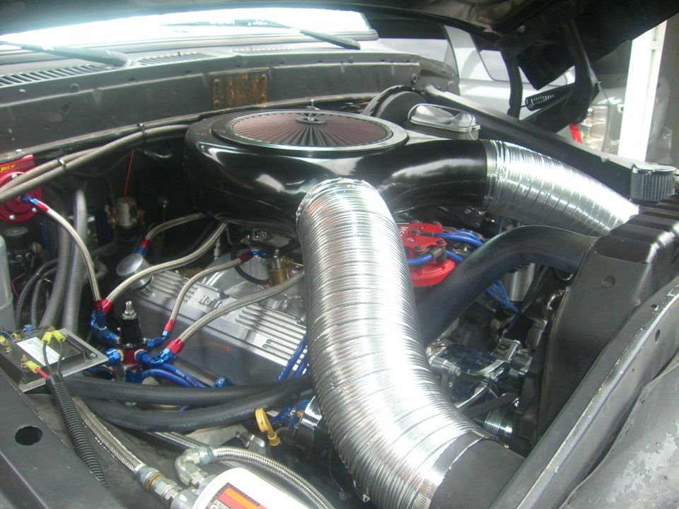 Engine bay view of a V8 engine with chrome valve covers, aftermarket air intake, and braided fuel lines