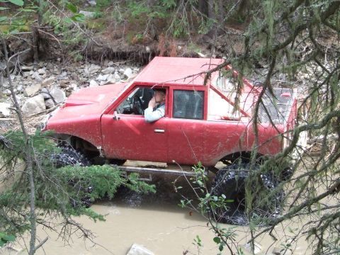1990 Ford Bronco - A modified red off-road vehicle navigates a muddy stream surrounded by trees and foliage.