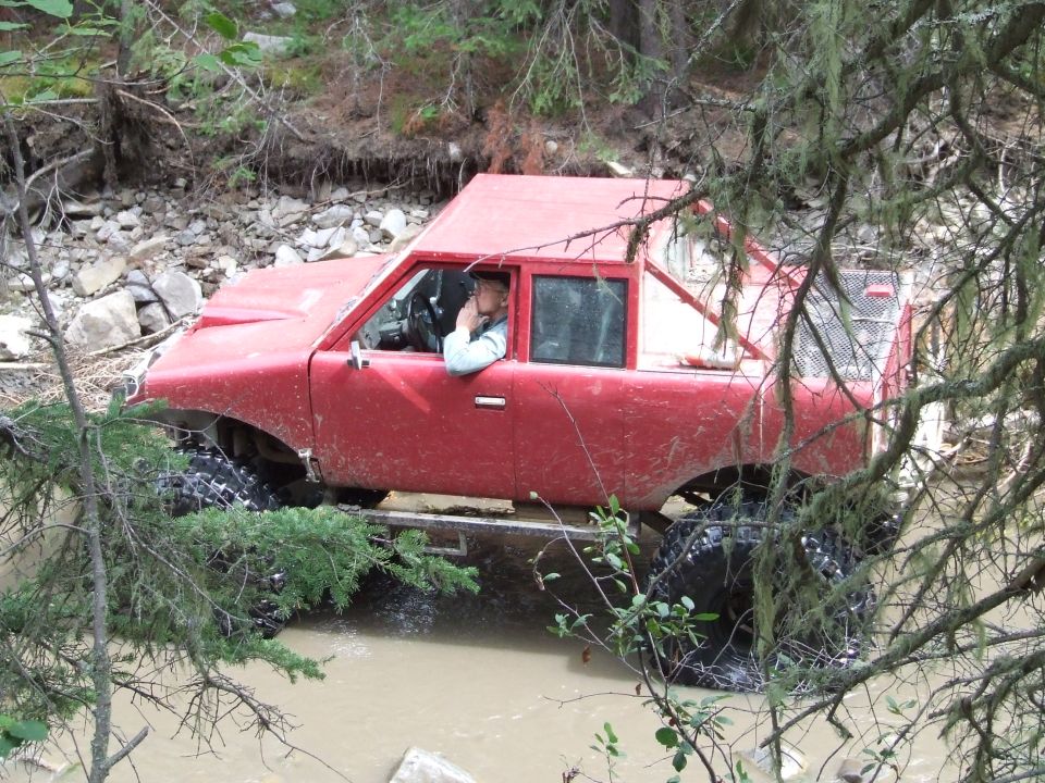 Red off-road vehicle with large tires driving through muddy water, driver visible inside