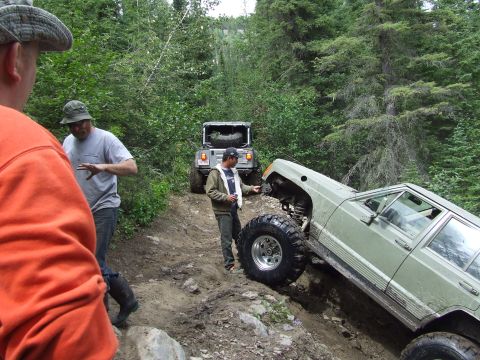 1990 Ford Bronco - Off-road vehicles tackle a challenging trail in a wooded area, with one vehicle at a precarious angle.